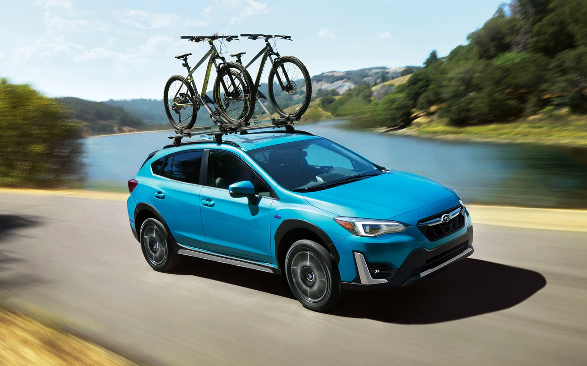 A blue Crosstrek Hybrid with two bicycles on its roof rack driving beside a river | Briggs Subaru of Topeka in Topeka KS