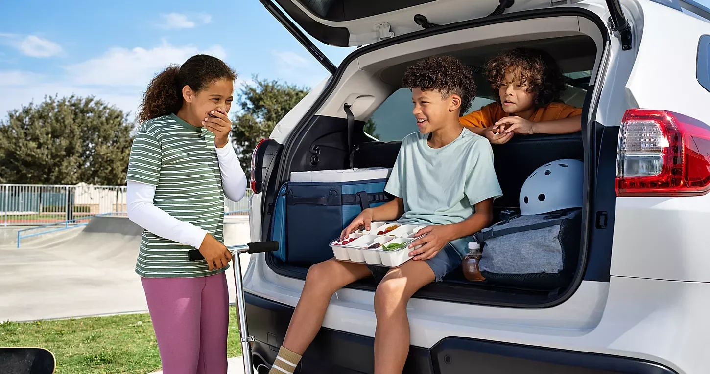 Two children sitting in the open boot of a Subaru while an adult stands beside it.