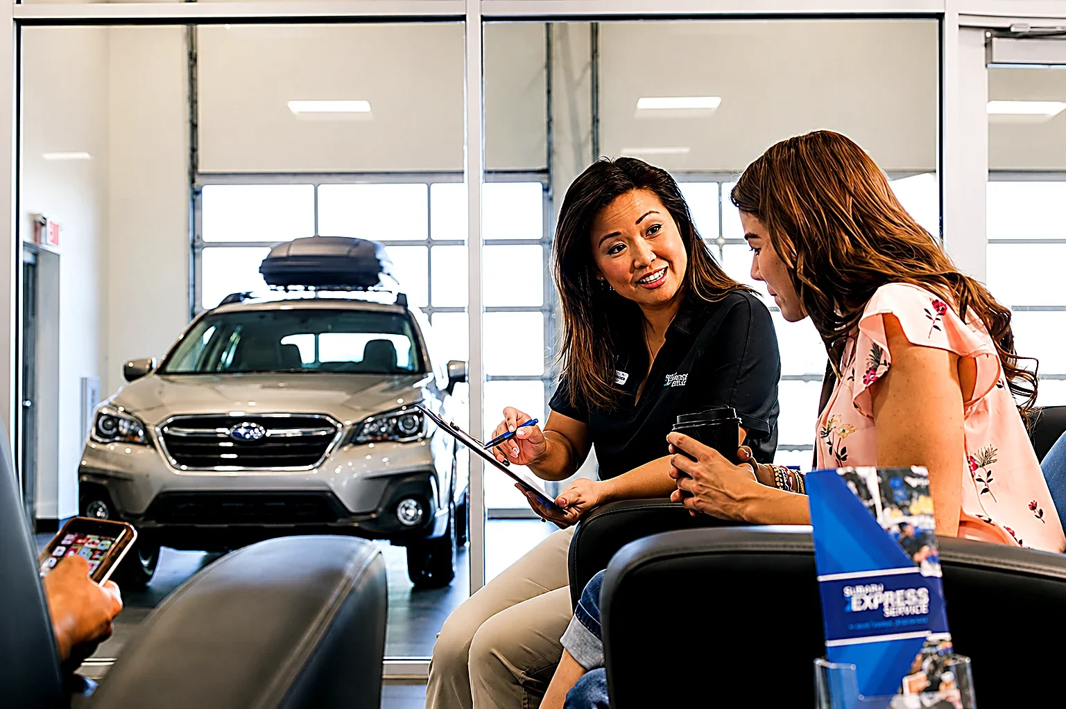 A Subaru advisor showing a tablet to a customer seated in a showroom, a vehicle behind them.