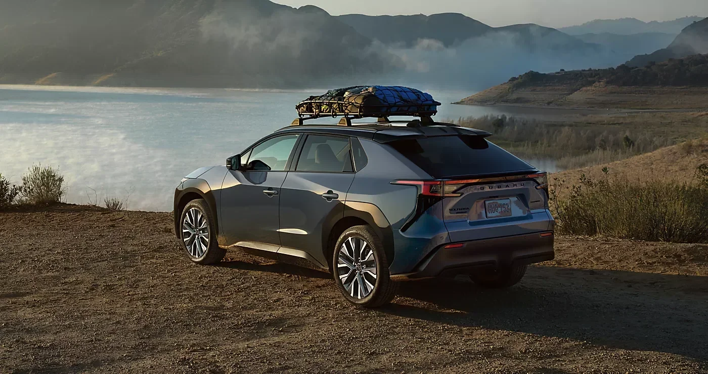 A Subaru Solterra parked on gravel by a lake, with hills behind it.