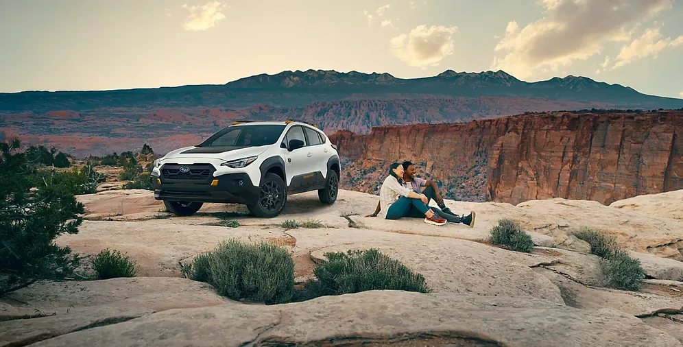 Couple resting on red rock beside a white 2026 Subaru Crosstrek at a canyon overlook