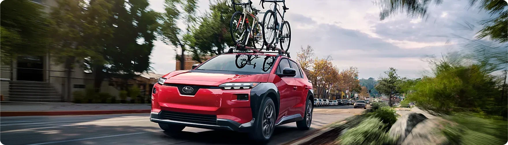 Red 2026 Subaru Solterra carrying bicycles on the roof through a tree-lined street